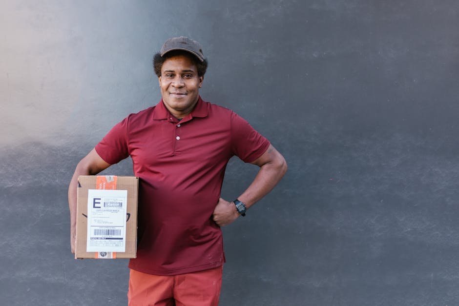 A smiling male delivery person wearing a maroon polo shirt, matching shorts, and a dark cap, standing against a plain grey wall. He holds a rectangular cardboard parcel with a shipping label and orang